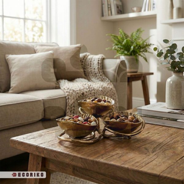 چیدمان آجیل و تنقلات داخل ظرف پذیرایی سه تکه طلایی روی میز جلو مبلی چوبی.